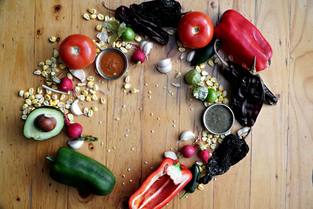 Flat lay arrangement of fresh Mexican cooking ingredients including tomatoes, bell peppers, avocados, corn, and chili peppers on a wooden table